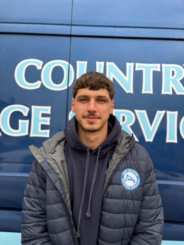 Person in navy quilted jacket with West Country Drainage Services logo, smiling, in front of company vehicle.